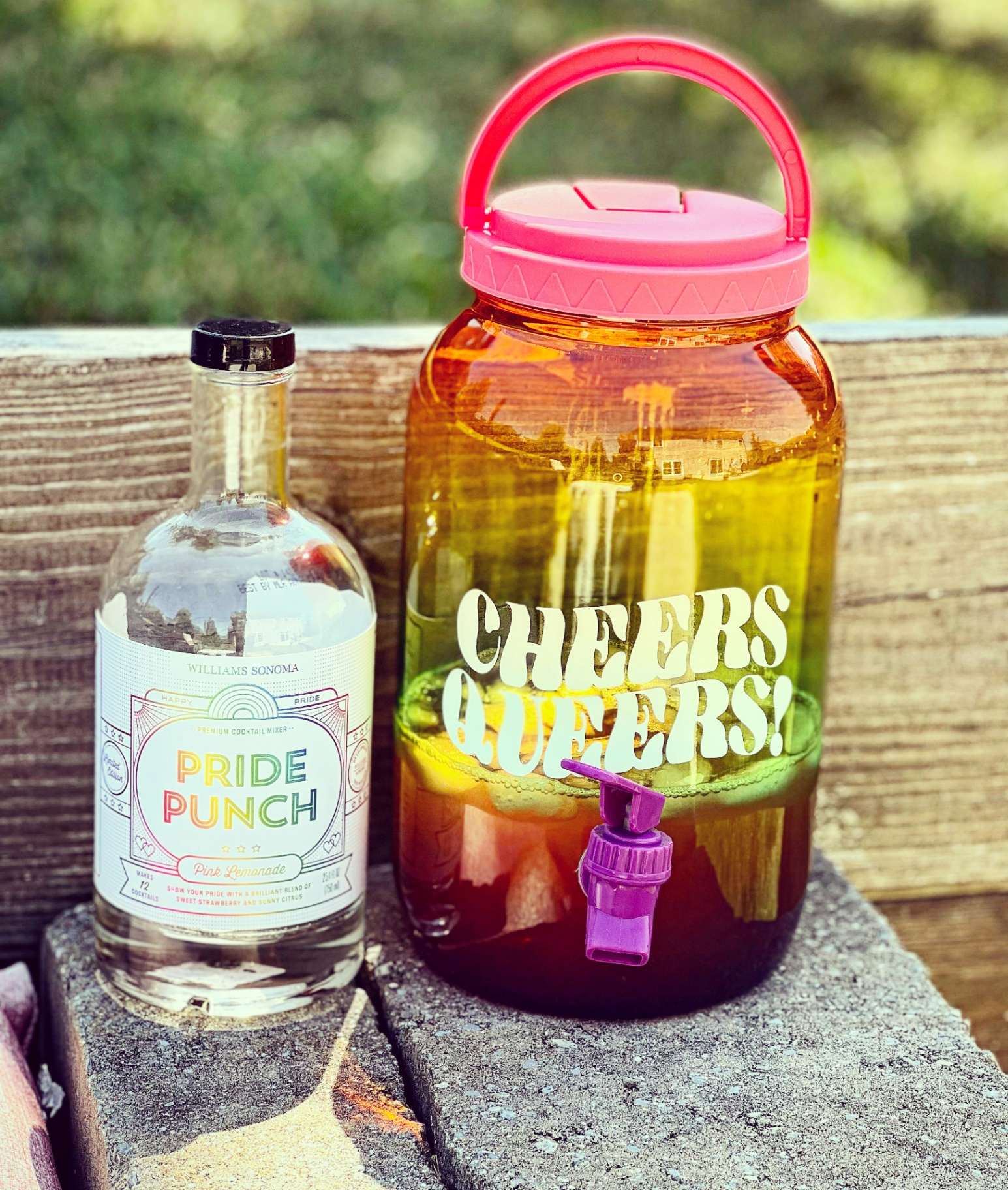 Product photography of a Williams Sonoma Pride Punch pink lemonade mixer beside a 'Cheers Queers' drink dispenser.